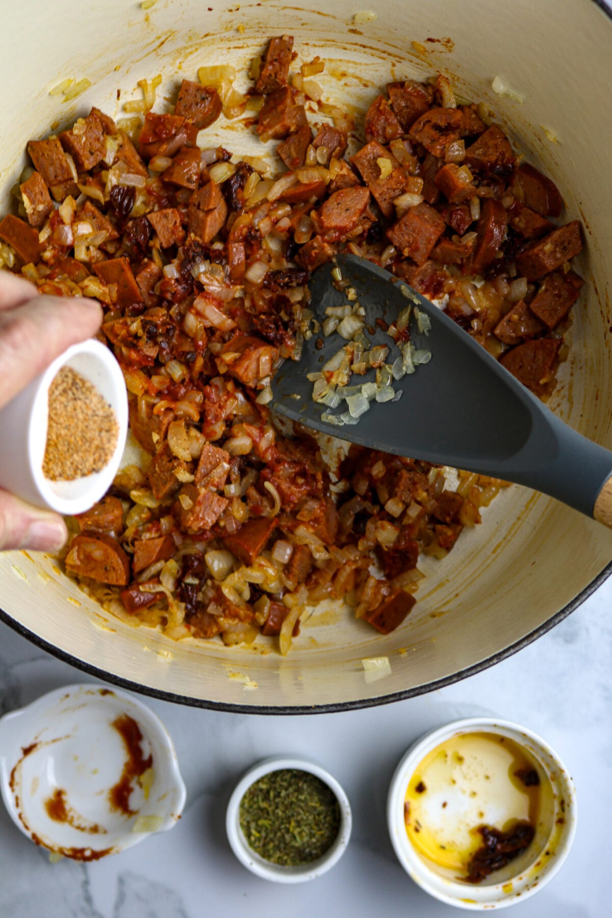 A pot of chopped sausage and a hand pouring seasoning over it.