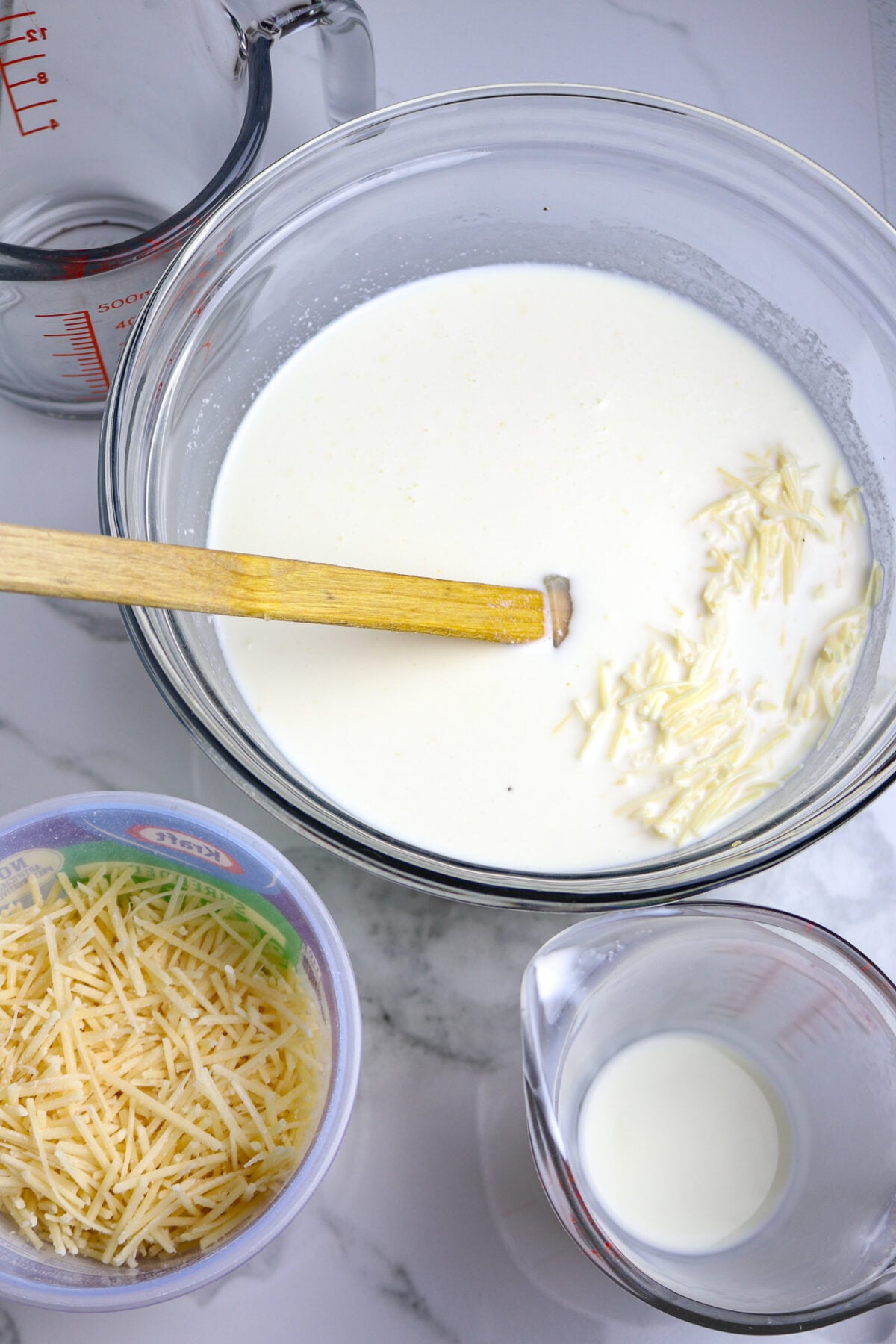 A wooden spoon in a glass bowl of cream and cheese next to a bowl of parmesan cheese and a2 empty measuring cups.