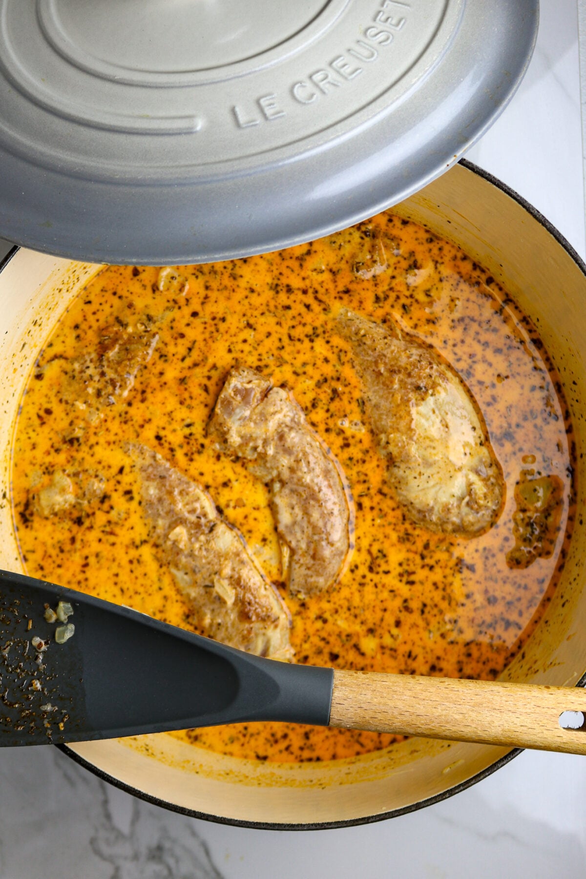 A gray Dutch oven filled with a golden sauce with chicken tenders and a spoon resting on top with the lid on the side.