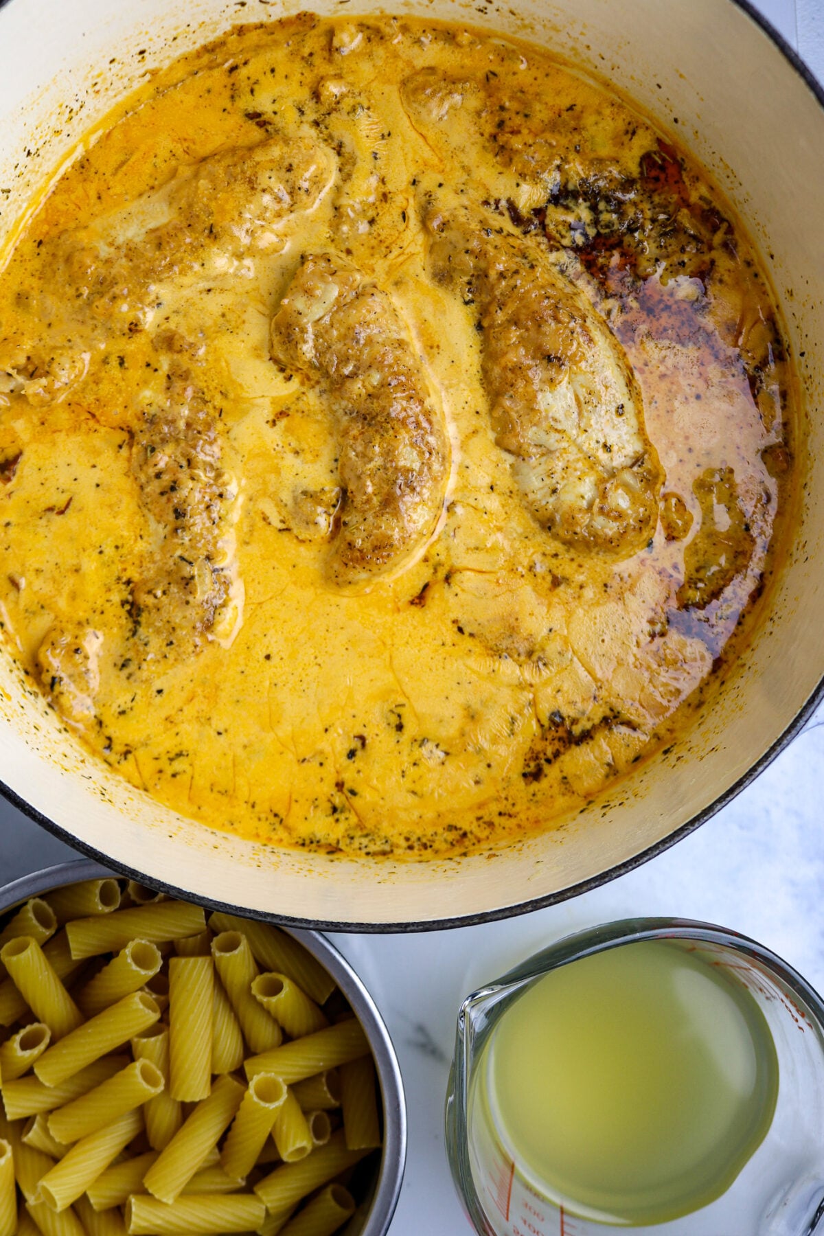 A golden broth for Marry Me Chicken in a large pot with chicken tenders next to a bowl of rigatoni pasta and a cup of chicken broth.