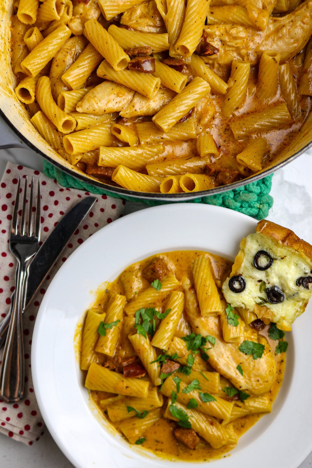 A white bowl of Marry me Chicken with a piece of cheesey bread next to it and placed next to a pot of the Pasta and chicken dish.