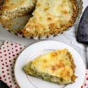 A whole cheese topped onion pie with a piece cut out and placed on a white plate.