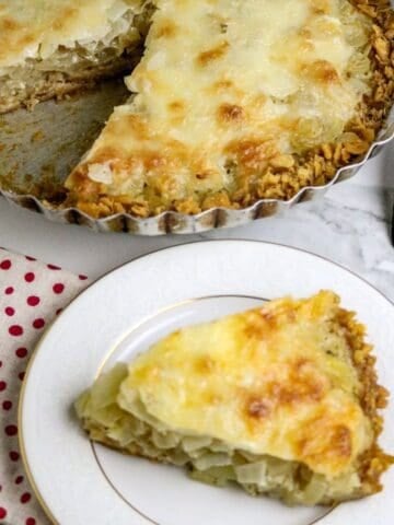 A whole cheese topped onion pie with a piece cut out and placed on a white plate.