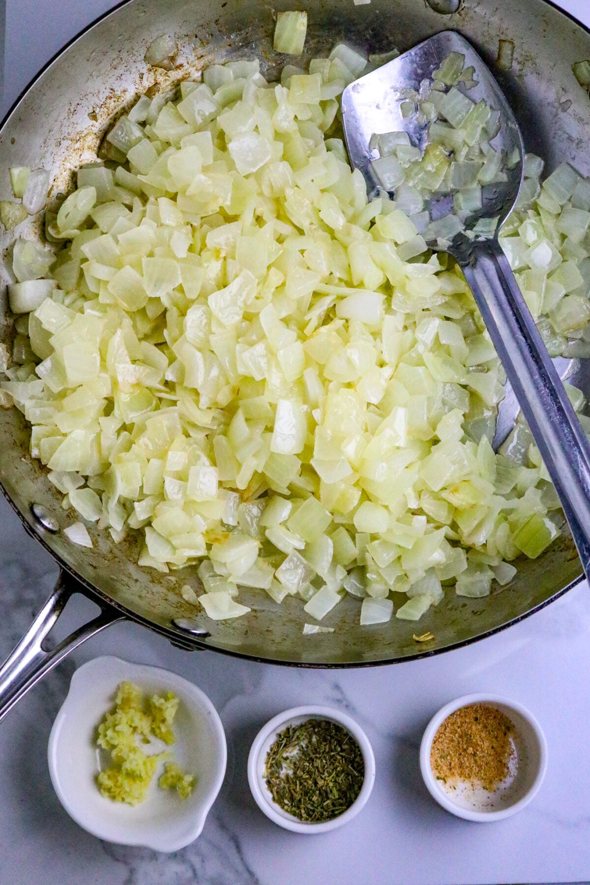 A silver skillet with chopped onions and a silver spoon and seasonings beside it.