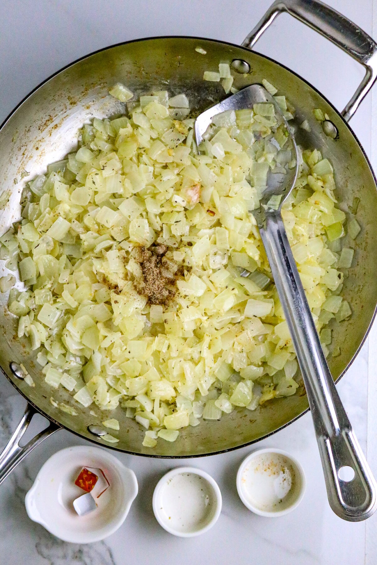 A silver skillet with chopped onions and a silver spoon and seasonings beside it.