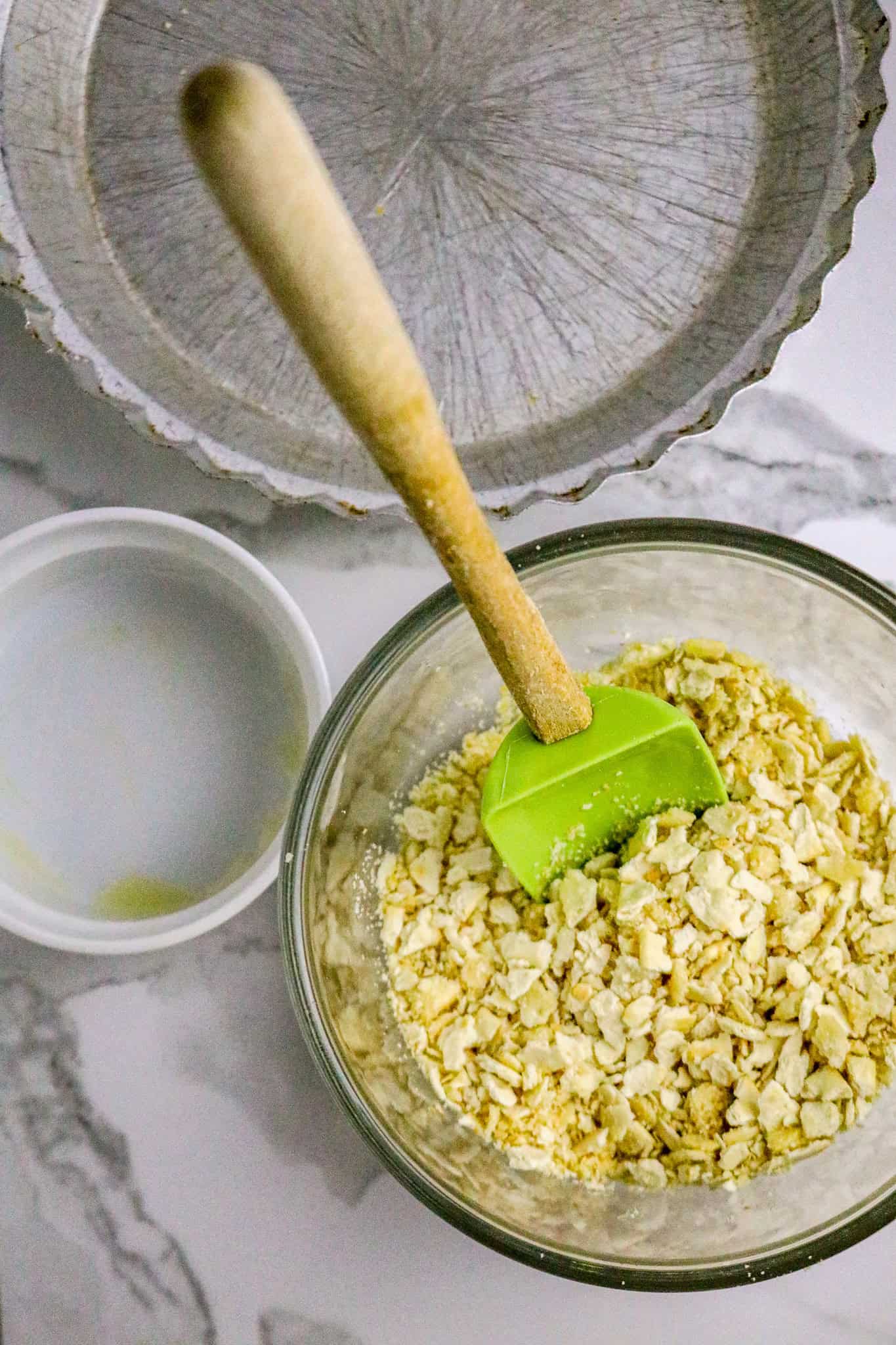 A glass bowl of cracker crumbs with a green spoonula in it next to an empty white bowl and a metal pie plate.