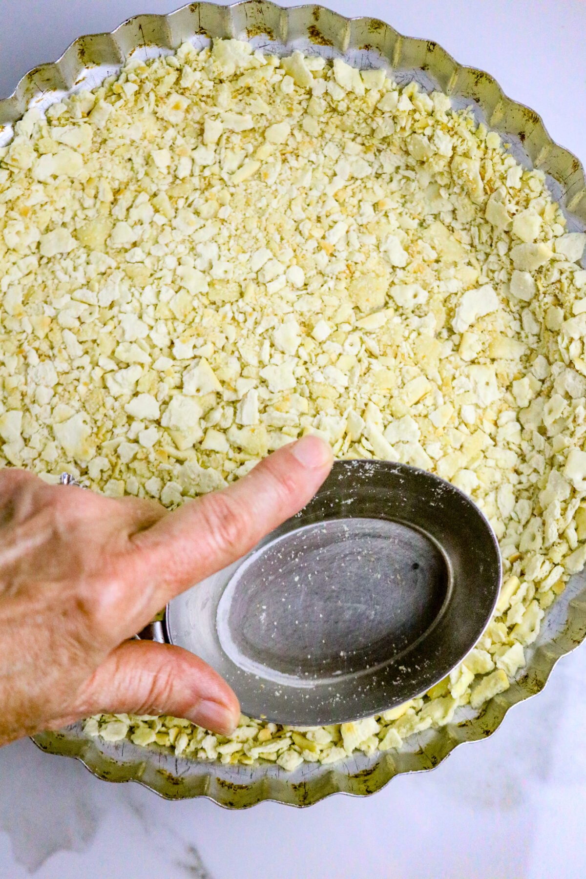 A metal pie plate with a cracker crumb crust and a hand pressing the pie crust into the pan with an oval metal measuring cup.