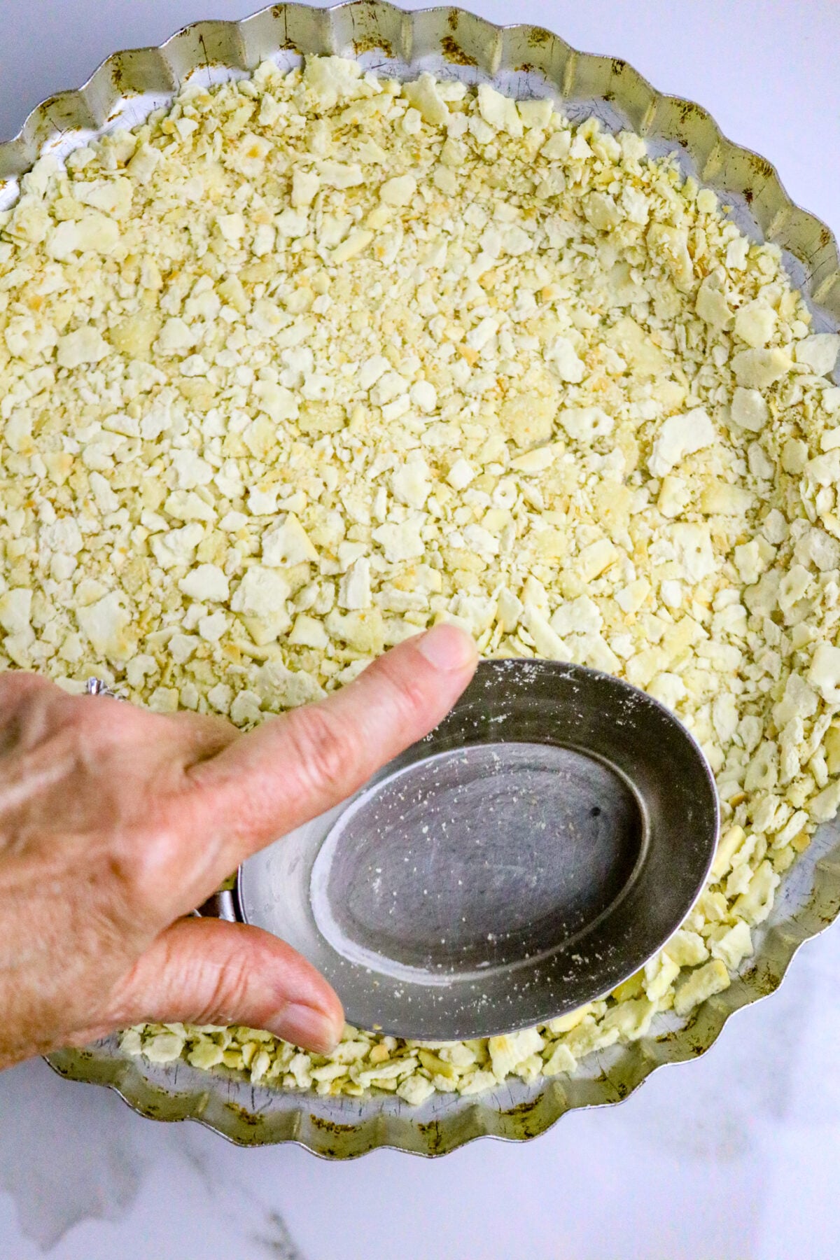 A pie plate lined with cracker crumbs and a hand pressing the edges with a silver measuring cup.