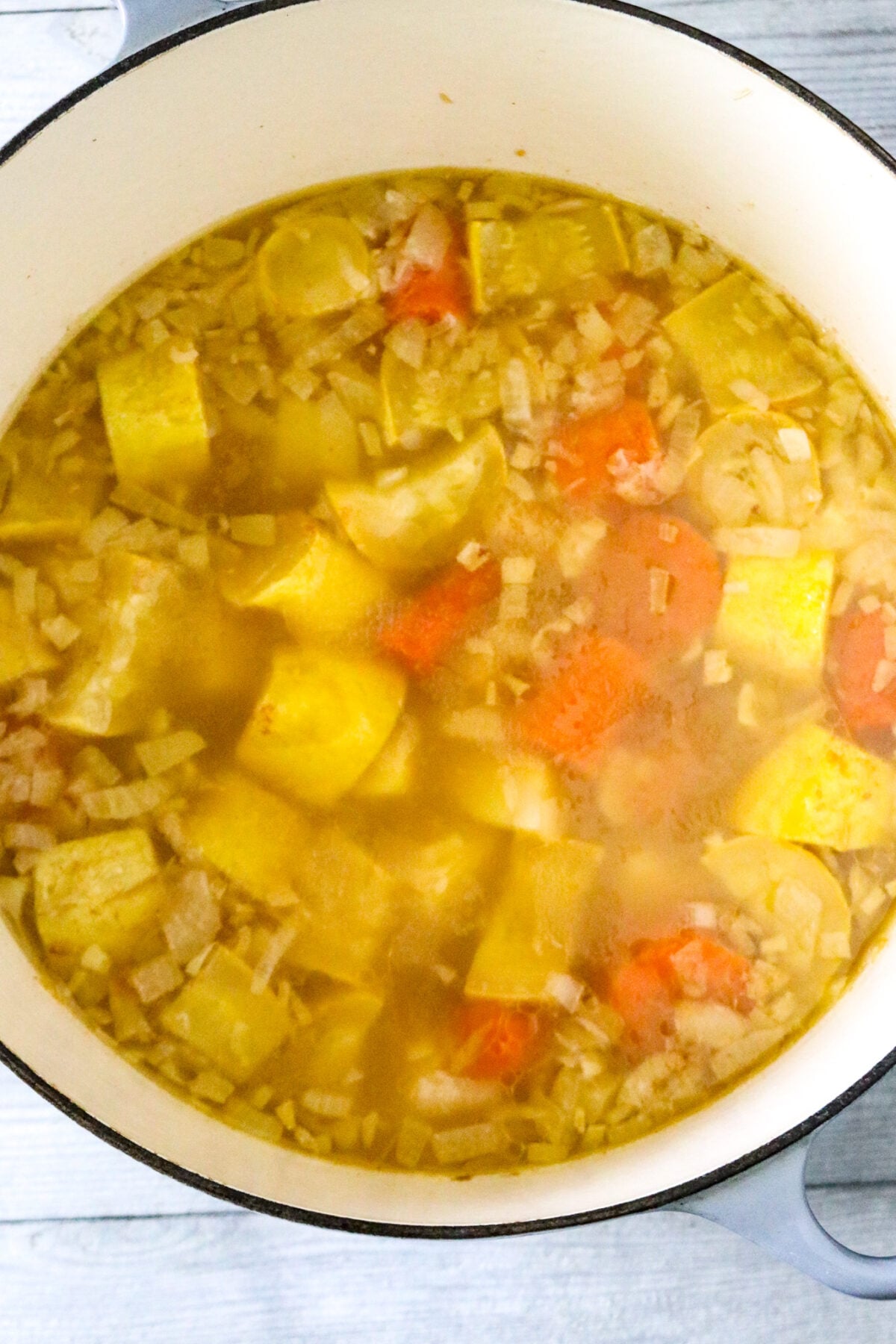 A Dutch oven pot with cooked chunks of carrots, potatoes, and squash in chicken broth with seasonings in it.
