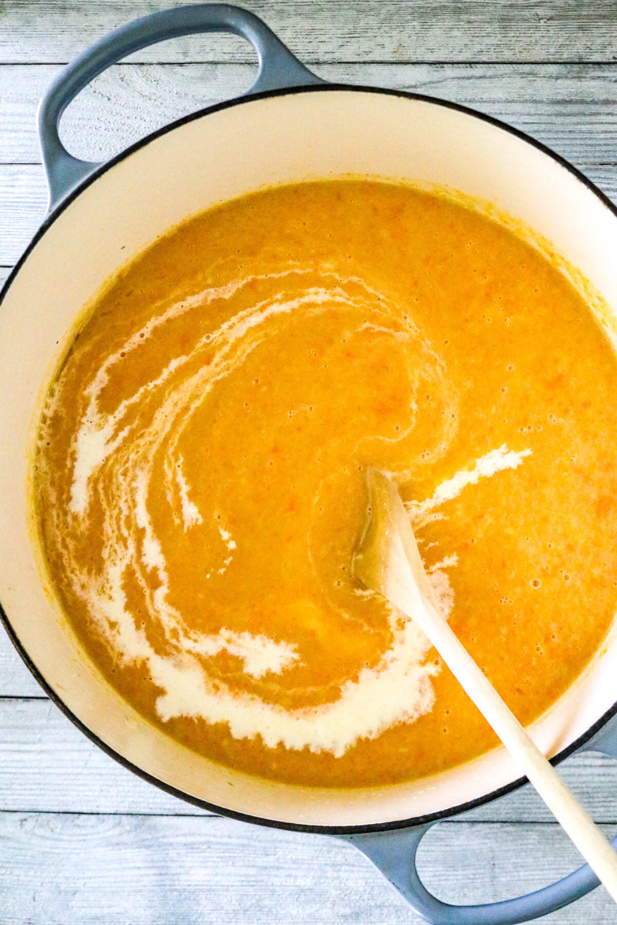 A Dutch oven pot with a squash soup and wooden spoon stirring cream into the soup.