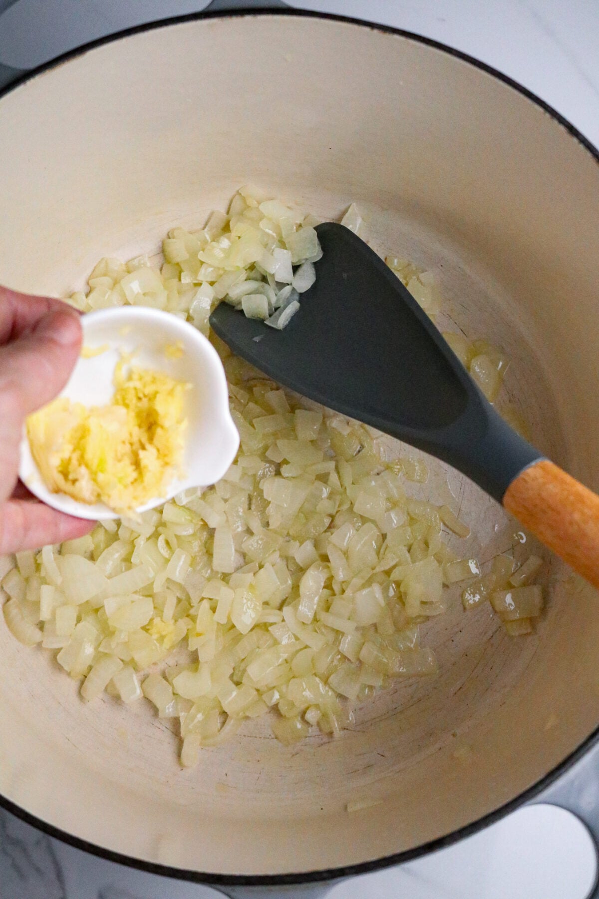 A hand pouring garlic from a small white bowl into a Dutch oven pot with cooked onions and a dark long-handled spoon