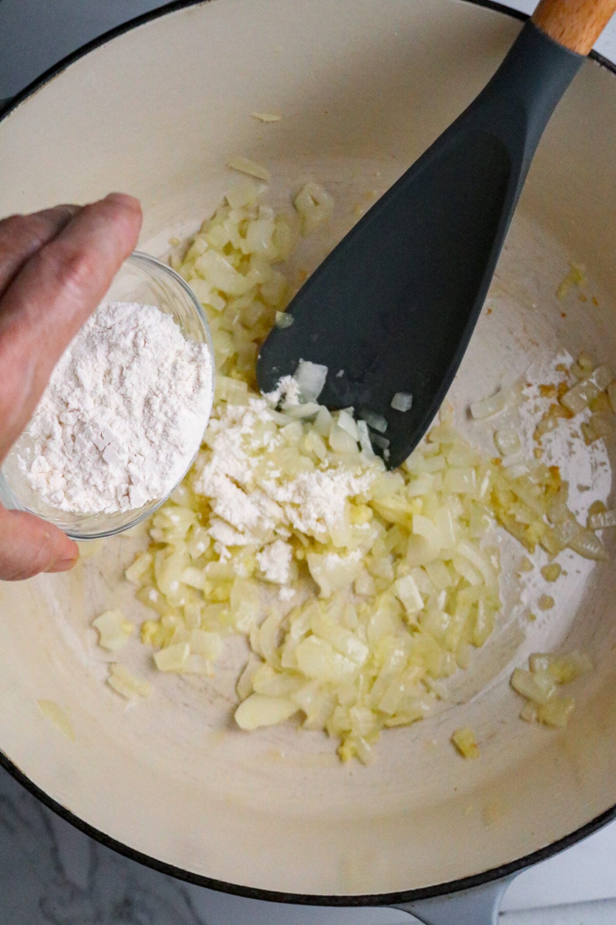 A hand pouring over flour from a small glass bowl into a Dutch oven pot with cooked onions and a dark long-handled spoon