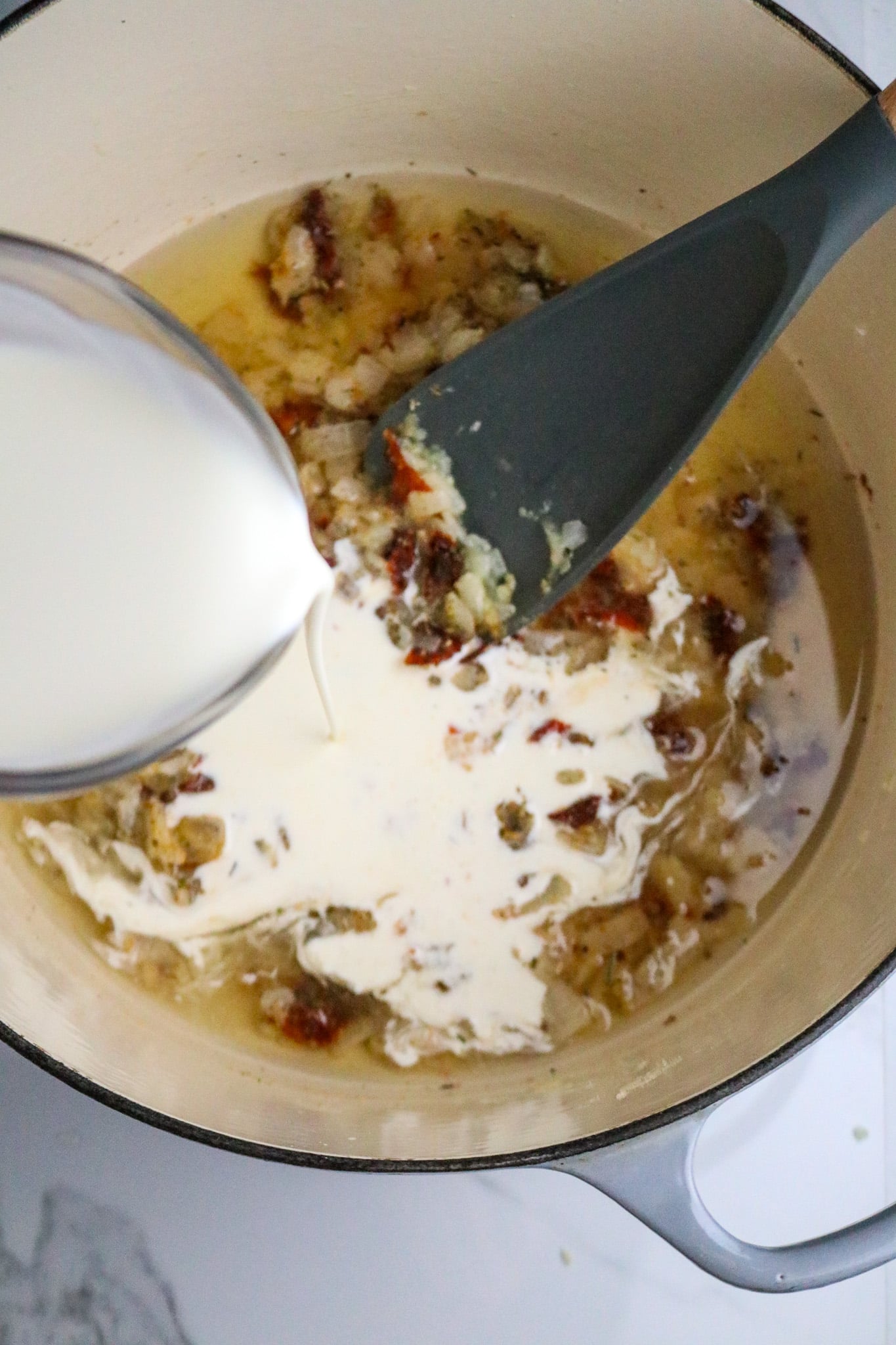 A cup of cream poured into a Dutch oven pot with a long-handled spoon in a clear sauce with seasonings and chopped vegetables.