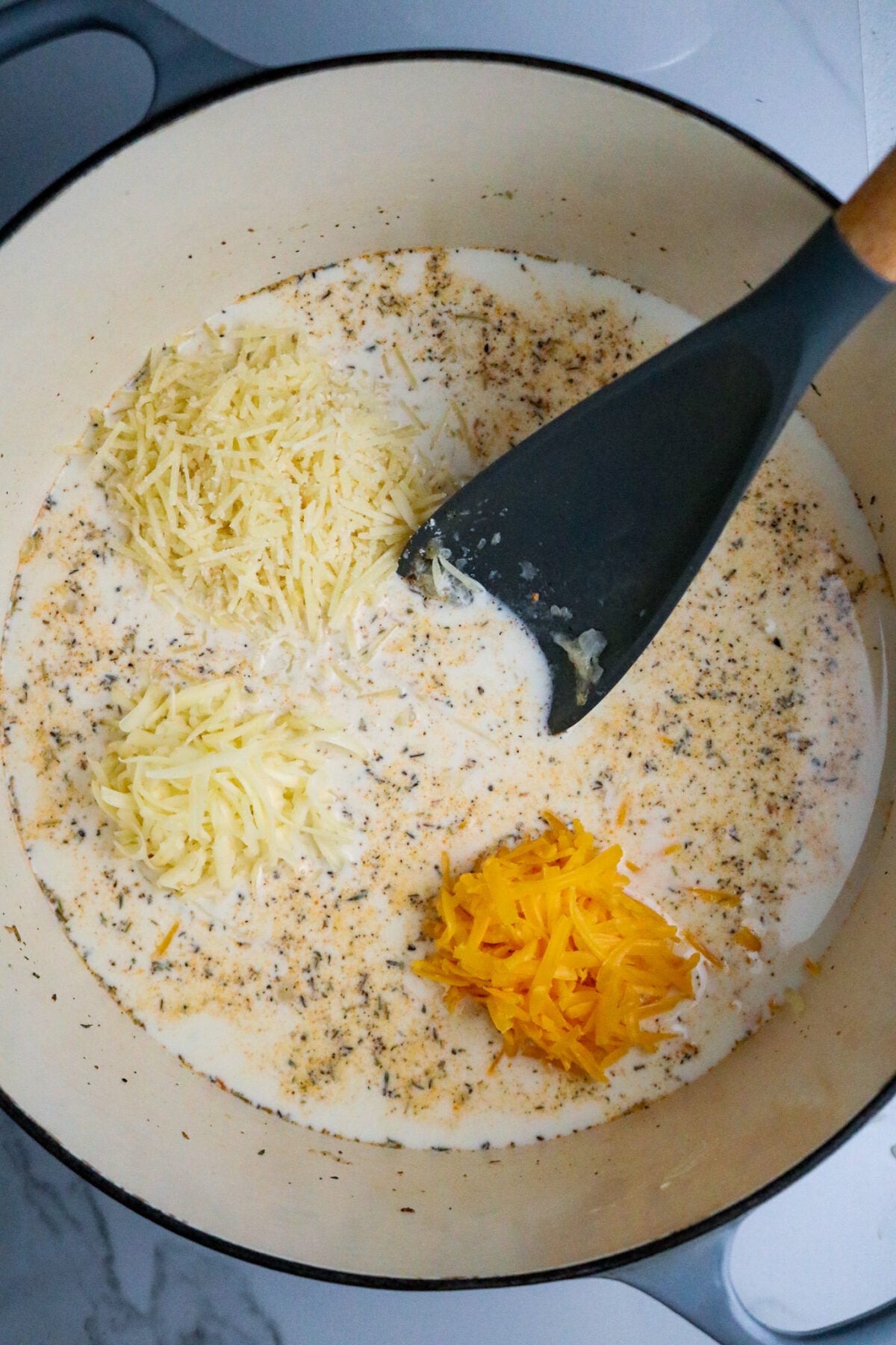 A dark long-handled spoon stirring in 3 mounds of grated cheese into a cream sauce.