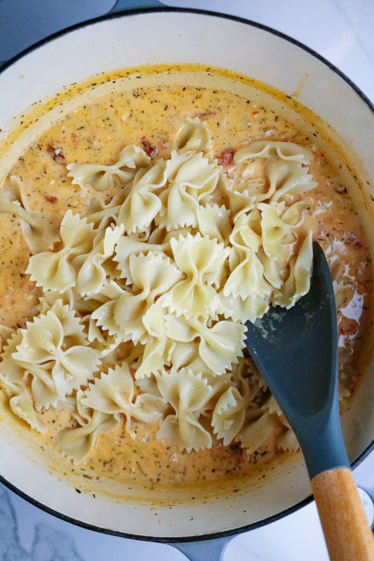A Dutch oven pot with a spoon stirring bowtie pasta into a cheesy cream sauce.