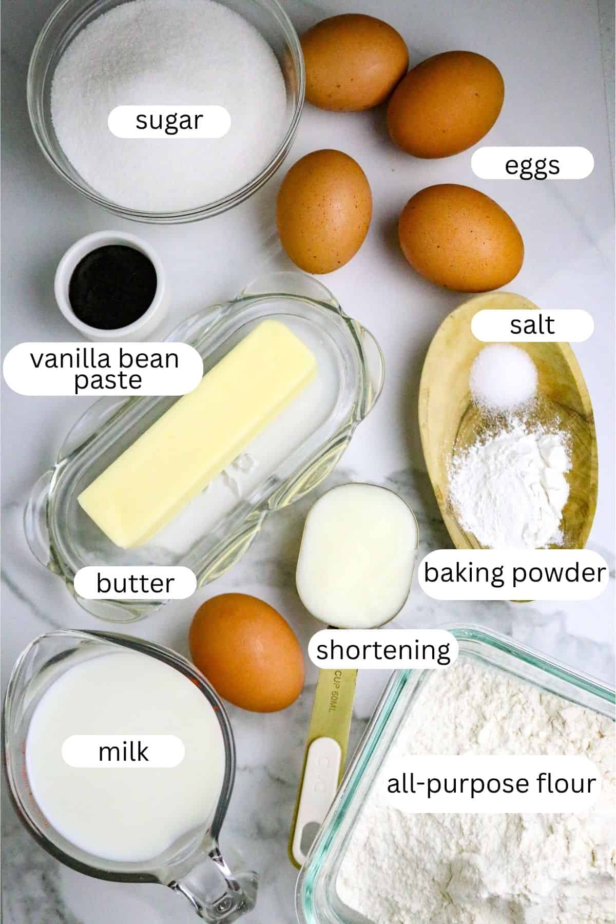 Ingredients of sugar, eggs, flour, milk, butter, shortening, salt, baking powder, and vanilla bean paste laid out on a white counter.