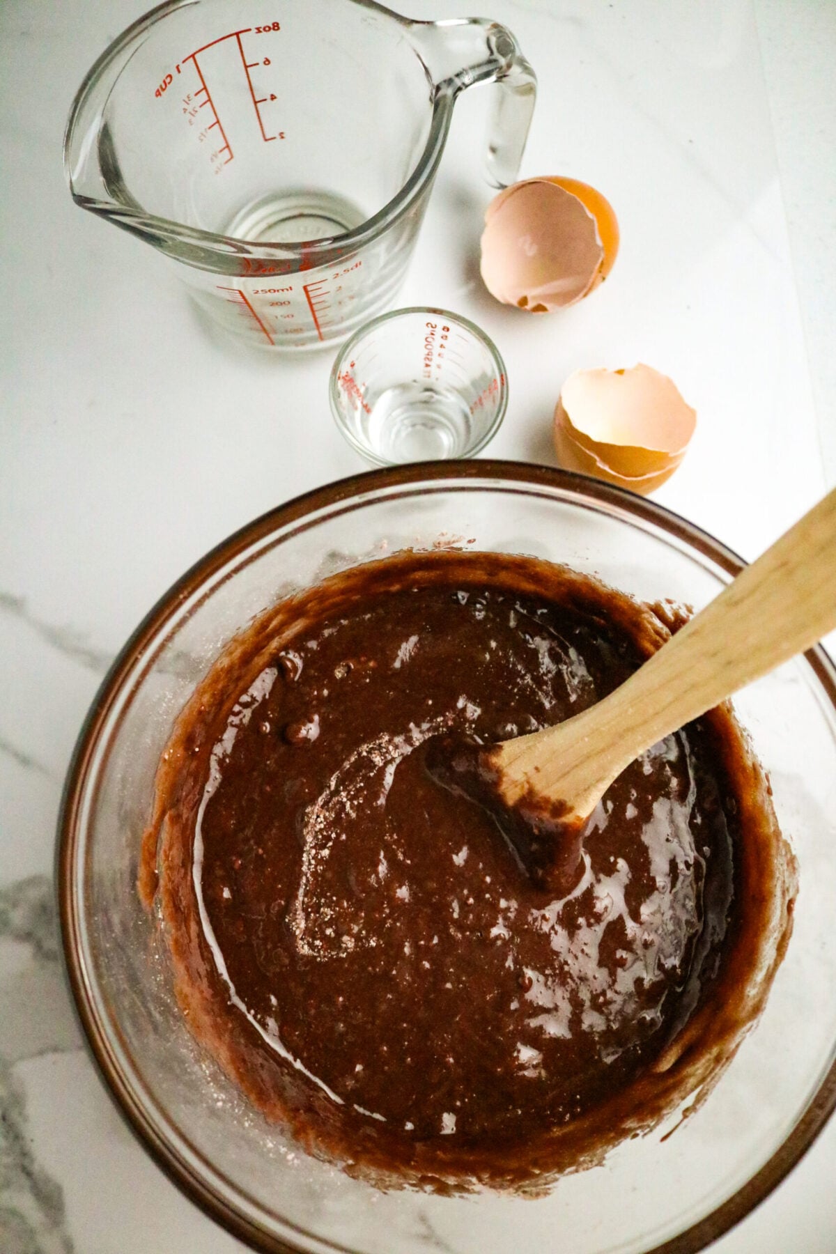 A glass bowl with a chocolate batter and a wooden spoon in it next to 2 broken eggs, a glass measuring cup, and a glass measuring shot glass.
