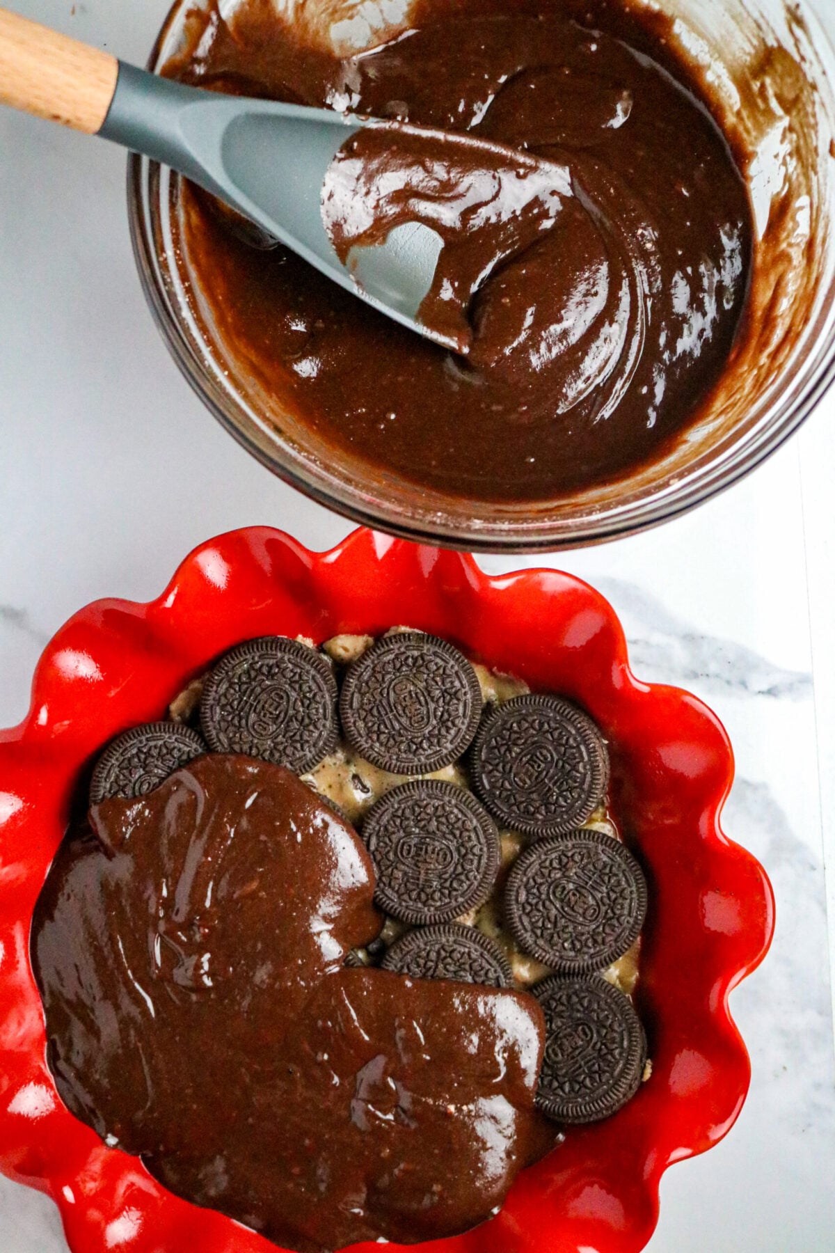 A glass bowl with a spoon in fudge batter next to a red pie pan with a layer of Oreo cookies and batter on half the top.