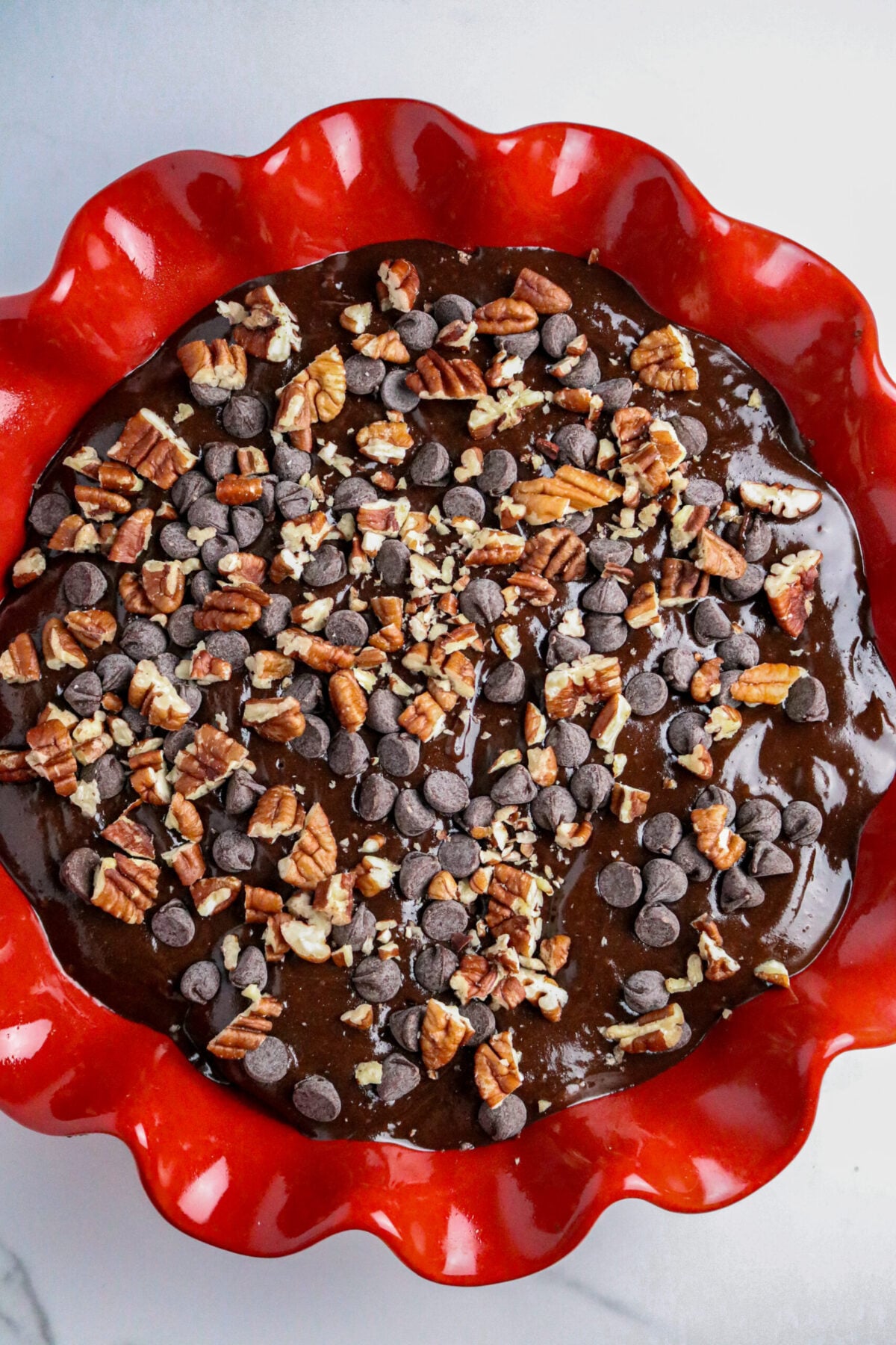 A red pie pan with fluted edges filled with a chocolate batter and topped with chocolate chips and chopped pecans.