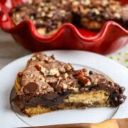 A red pie plate with a brownie pie inside covered with nuts and chocolate chips and a slice of pie on a white plate.