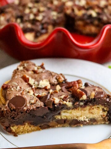 A red pie plate with a brownie pie inside covered with nuts and chocolate chips and a slice of pie on a white plate.